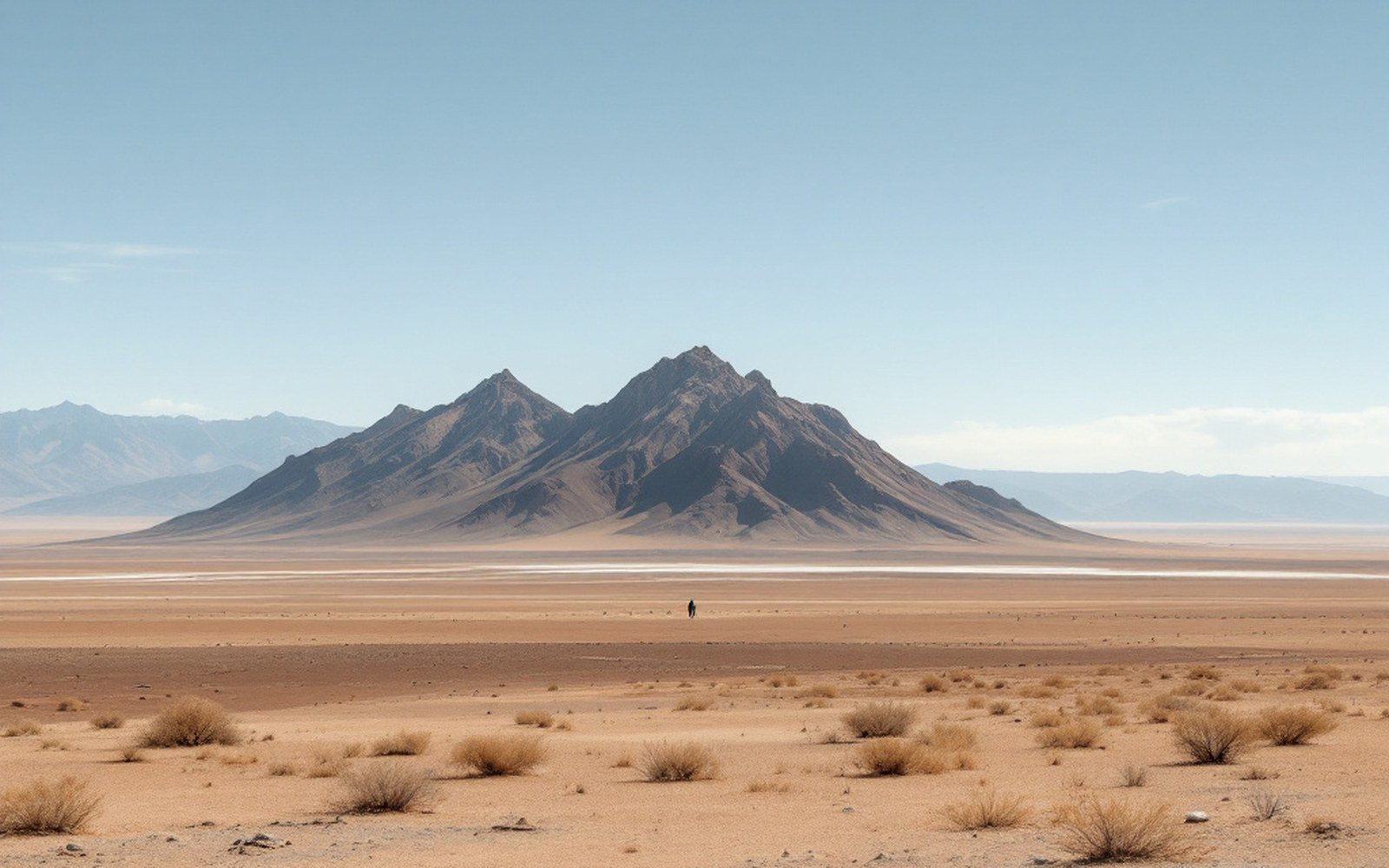 A lone figure walks across a vast desert plain beneath a jagged peak