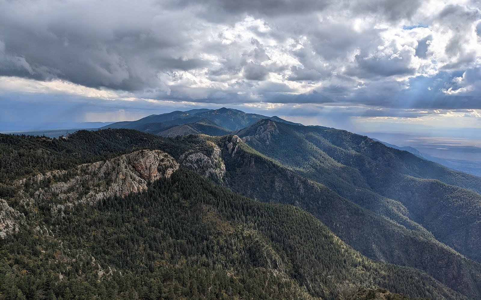 Forested ridgeline of the Manzano Mountains under a dramatic cloudy sky with sunbeams